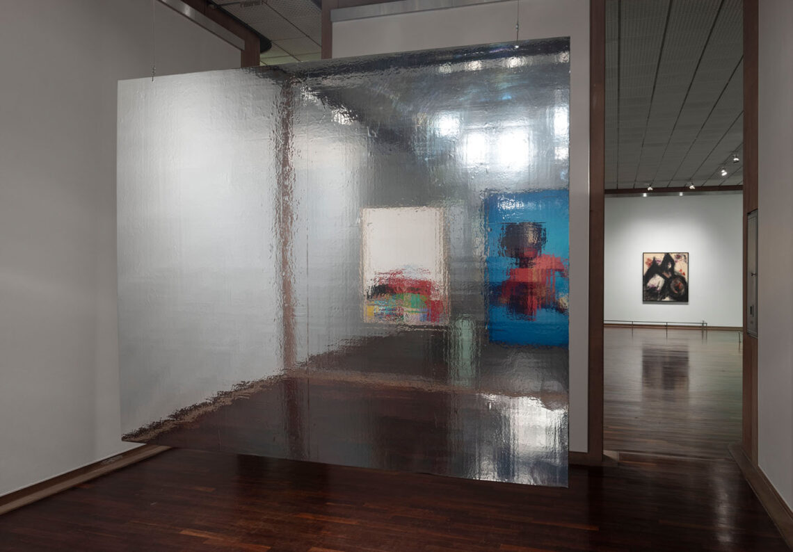 Two works of art on the wall opposite are reflected by a silver foil surface hanging freely in the room. On the left, a rather white picture is blurred, on the right a slightly larger one with a doll dressed in red with a dark head. The room otherwise has parquet flooring and white walls. In the background, an almost square painting with dark structures against a light background can be seen on a white wall.