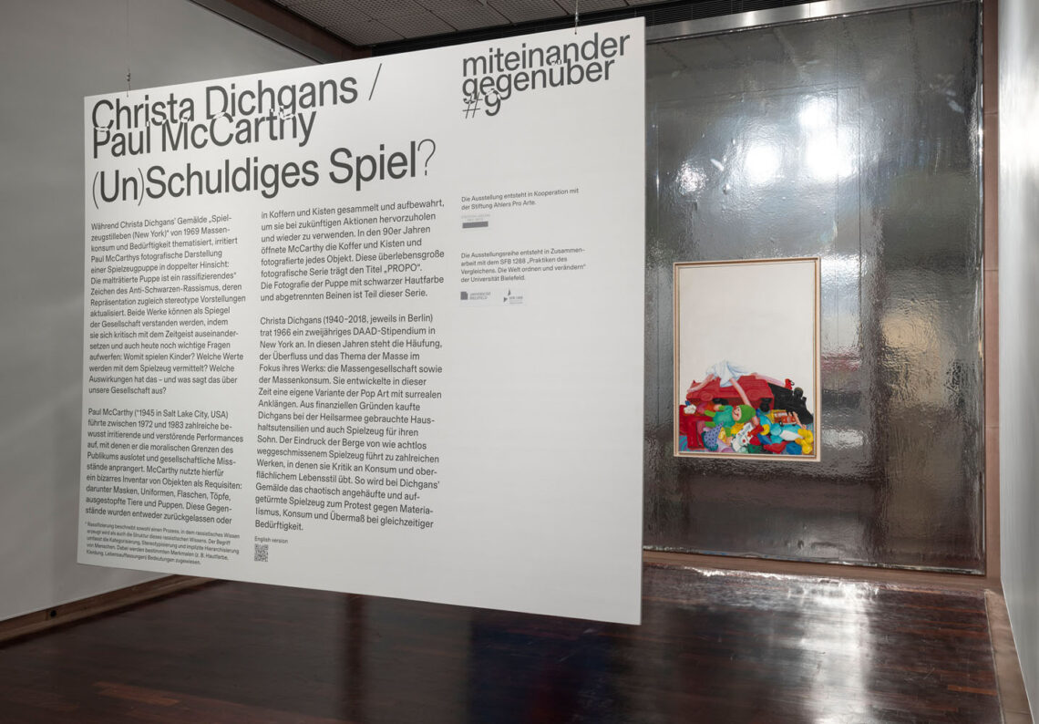 On a wall with reflective silver foil in the background is a painting with a pile of toys against a white background: dolls, stuffed animals, cars, with a doll with long limbs and a white dress lying on top. In the foreground of the photo is a text hanging in the room with explanations about this small exhibition.