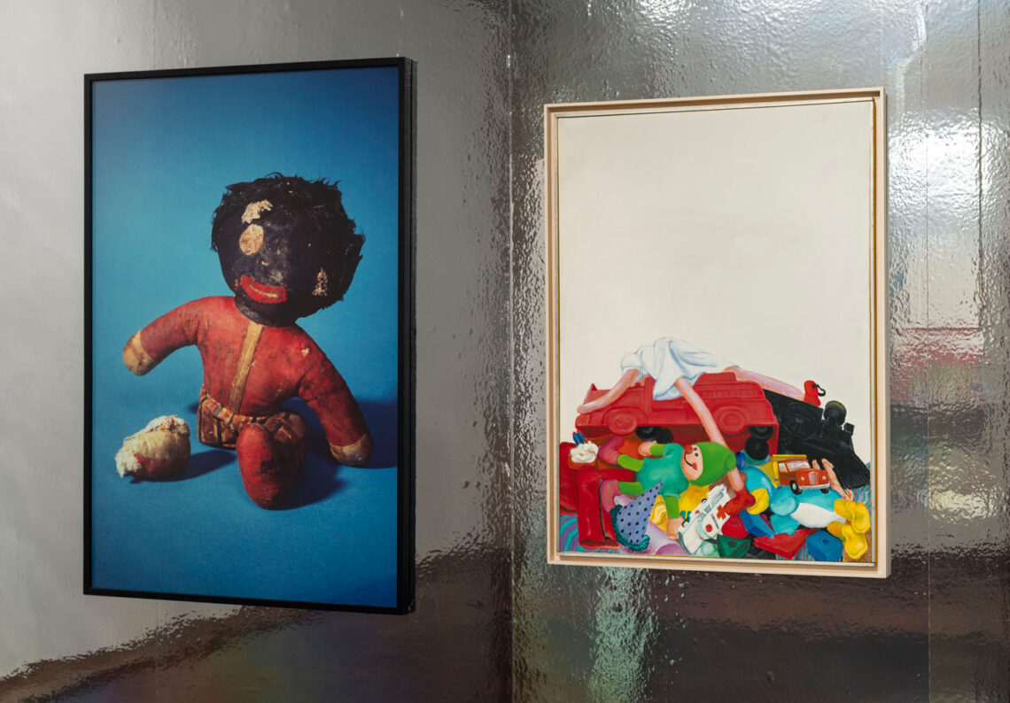 Two pictures hang on a wall with reflective silver foil. On the left is a photo of an old doll with a black head and thick lips. The doll is broken: the right leg lies severed in front of her. Behind her is a dark blue background. On the right, a painting with a pile of toys against a white background: dolls, stuffed animals, cars, on top of which lies a doll with long limbs and a white dress.