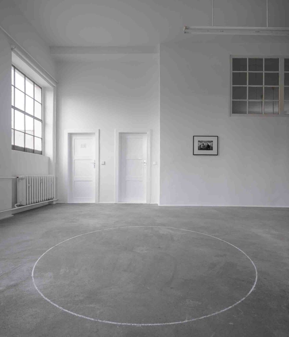 An empty room with high white walls and a dark floor. A large white circle has been drawn on the floor with chalk. Windows and doors can be seen in the background and a black framed photograph hangs on the wall